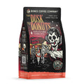 The front of a 12 ounce bag of Bones Coffee Company From Dusk til Dawn coffee. Its flavor is jelly donut, and it has a skeleton dressed like a vampire holding a jelly donut while people leave a donut shop on the art.