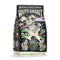 The front of a 12 ounce bag of Bones Coffee Company White Rabbit flavored coffee. It has a skeleton being carried by a white chocolate rabbit through a field of easter eggs on the art.