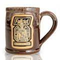 The front of the Bones Coffee Company Ghoul hand thrown mug with the Wasteland Crunch art on the golden medallion. It is inspired by Zenimax and Amazon’s Fallout show. The mug is chocolate colored with a sand white glaze on top.