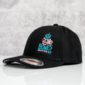 A black hat on top of a marble surface. The Bones Coffee Company logo is on the hate and is colored blue and pink. It has a skull that looks like a mug with steam coming out of the top of it. A sticker on the brim of the cap says Flexfit.